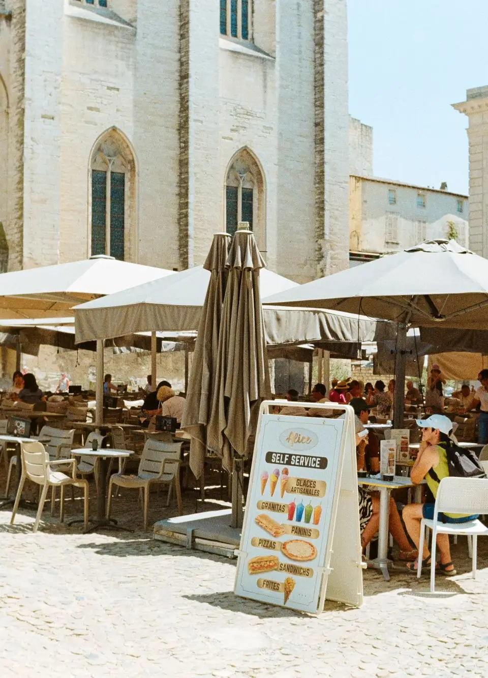 Lively summer scene at an outdoor cafe in Avignon, France, with people dining under umbrellas.