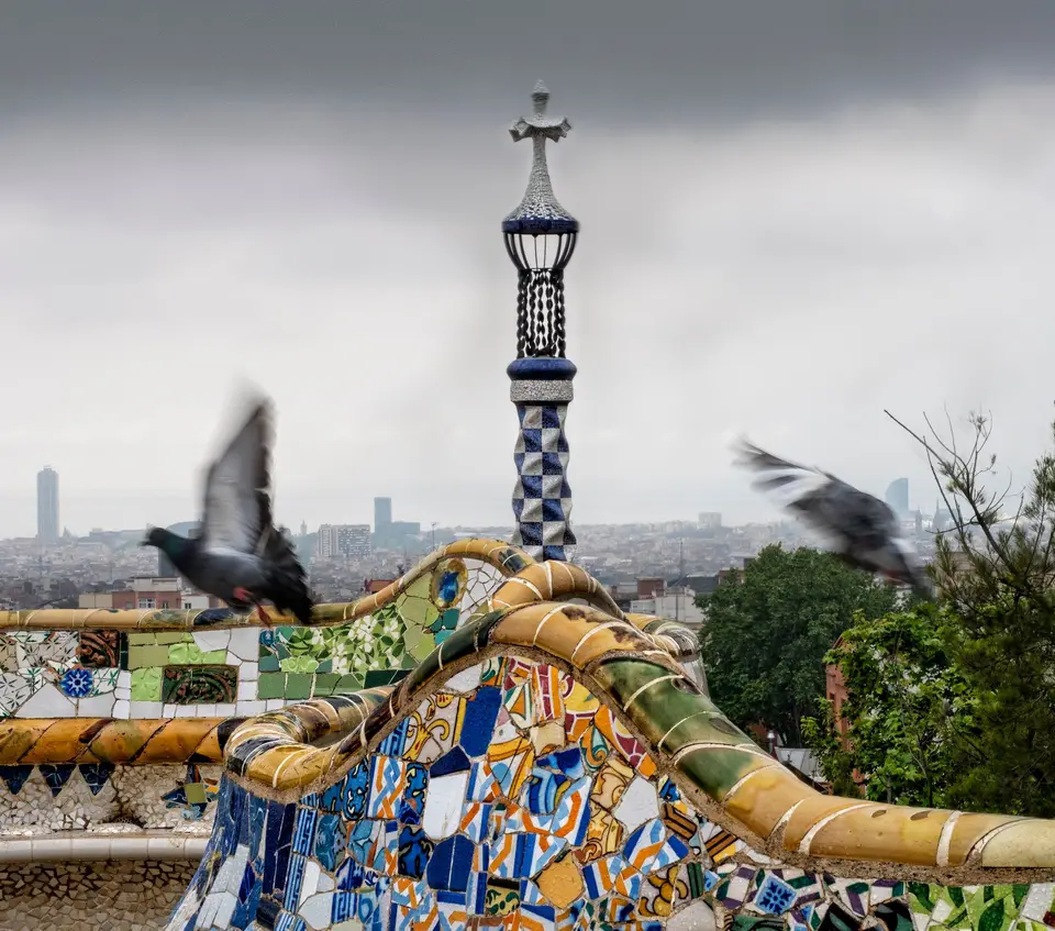 Vibrant mosaic with flying pigeons at Park Güell, Barcelona, under a cloudy sky.