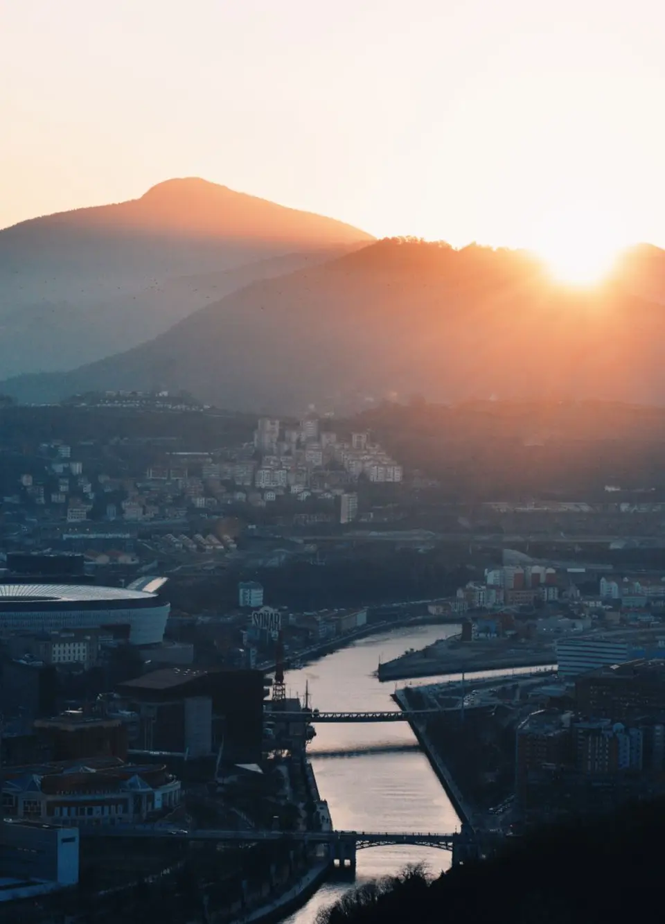 A breathtaking aerial view of Bilbao with sunset casting a warm glow over the city and surrounding hills.