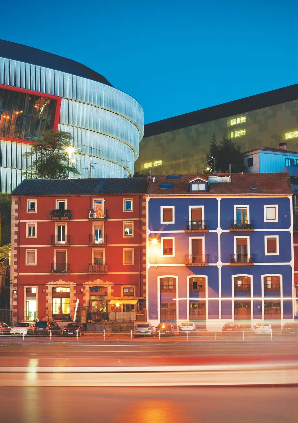 Colorful riverside buildings and San Mamés Stadium in Bilbao illuminated against the night sky.