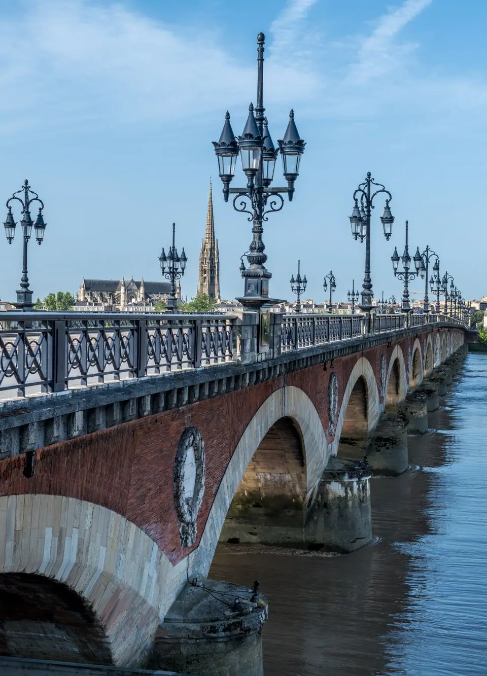 Scenic view of the iconic Pont de Pierre bridge in Bordeaux under a blue sky, reflecting on the calm waters of the Garonne River.