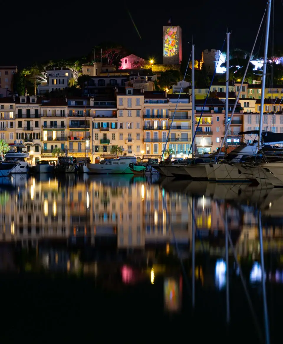 Beautiful night view of Cannes harbor with yachts and illuminated buildings reflecting in the water.