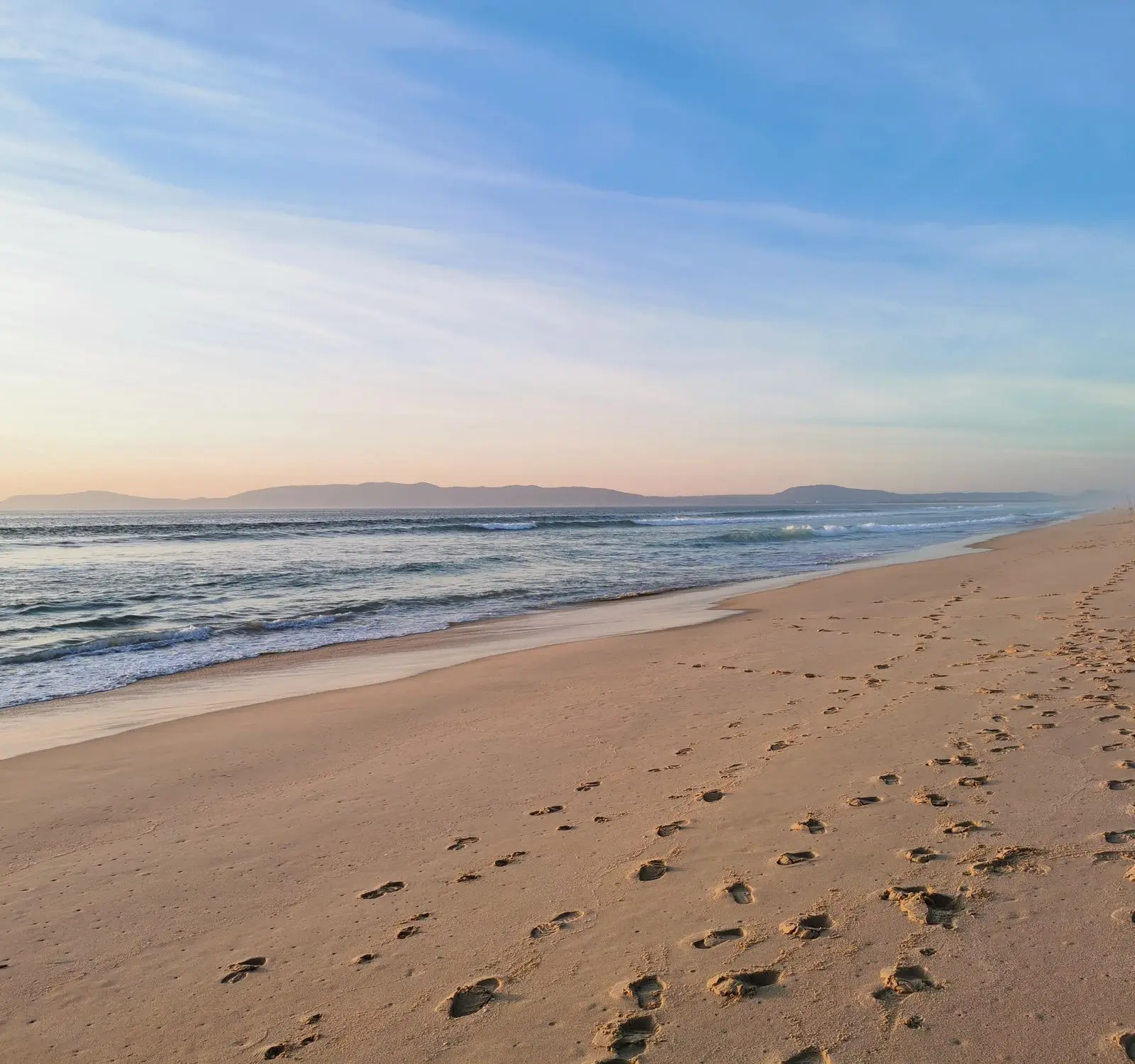 Peaceful sandy shore with footprints and gentle waves at sunset in Comporta, Portugal.