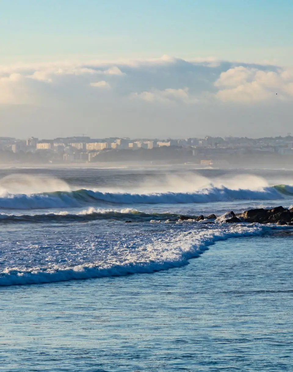 A surfer approaches ocean waves at Costa da Caparica beach with a clear sky backdrop.