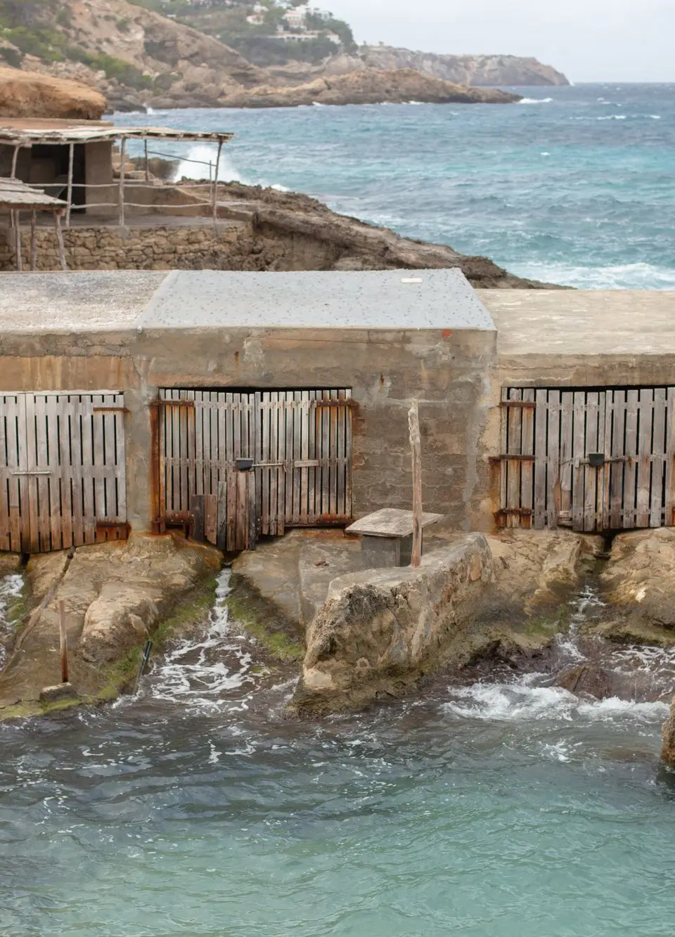 Rustic boathouses on Cala Tarida's Mediterranean coast in Ibiza, Spain, showcasing rocky terrain.