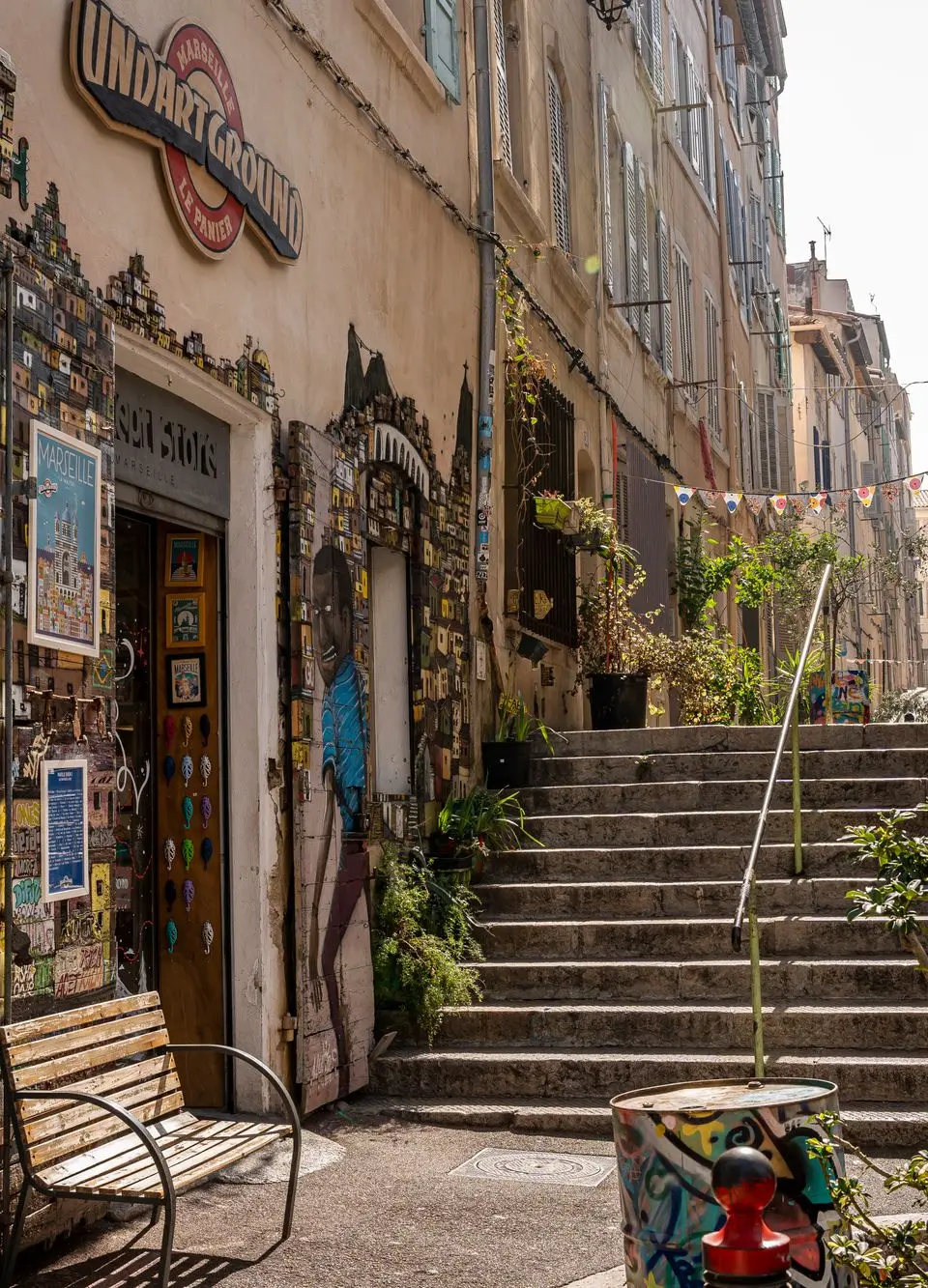 Sunlit narrow street in Marseille adorned with vibrant murals and lush plants on a bright day.