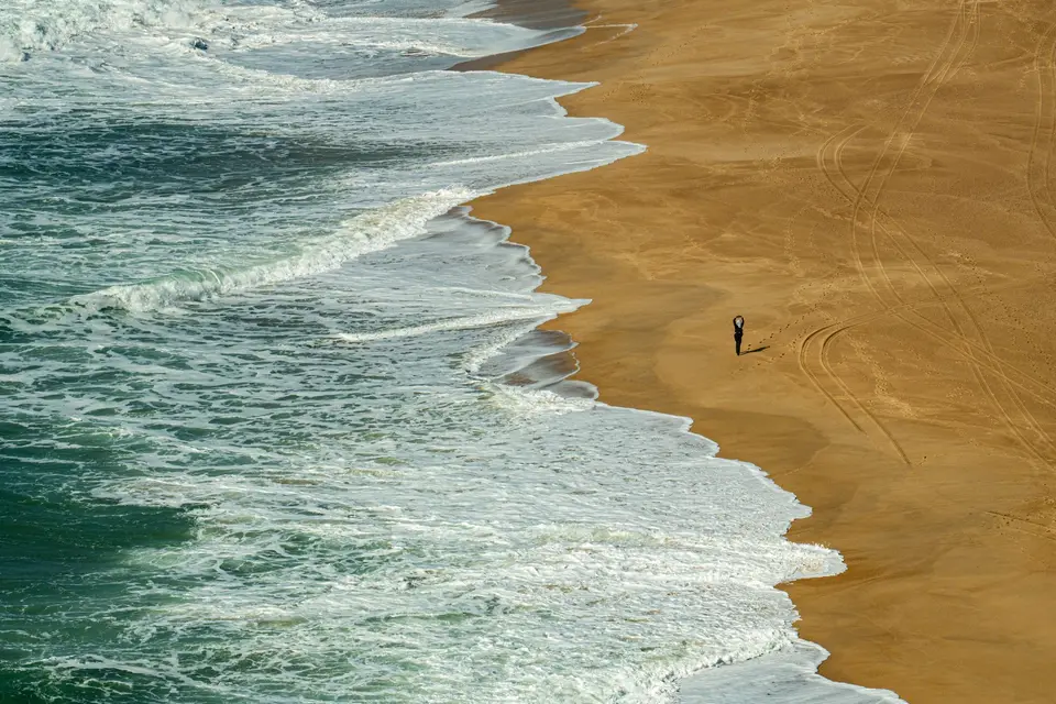 A lone person stands on the expansive sandy shores of Nazaré, Portugal with waves crashing nearby.