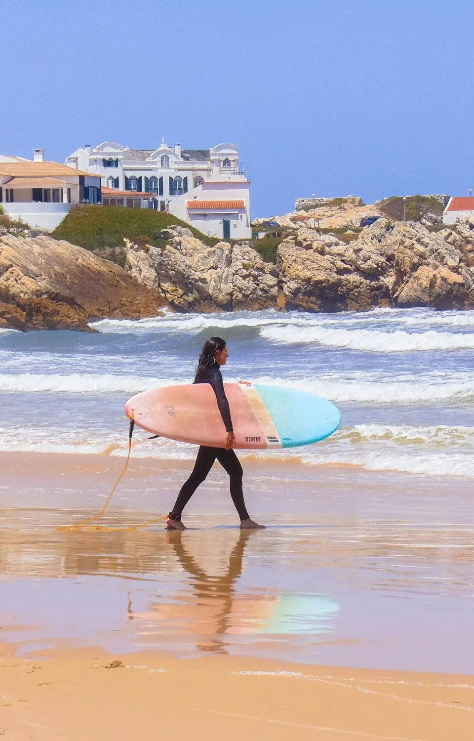 A surfer carrying a colorful surfboard strolls along the scenic beach in Peniche, Portugal.