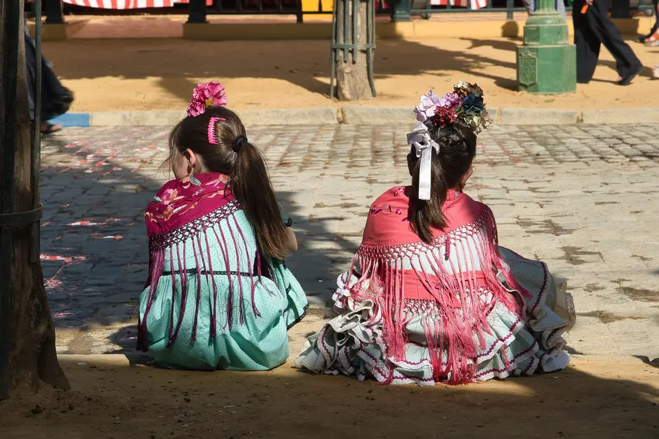 Girls in traditional flamenco dresses sitting on sidewalks during Seville's festive Feria.