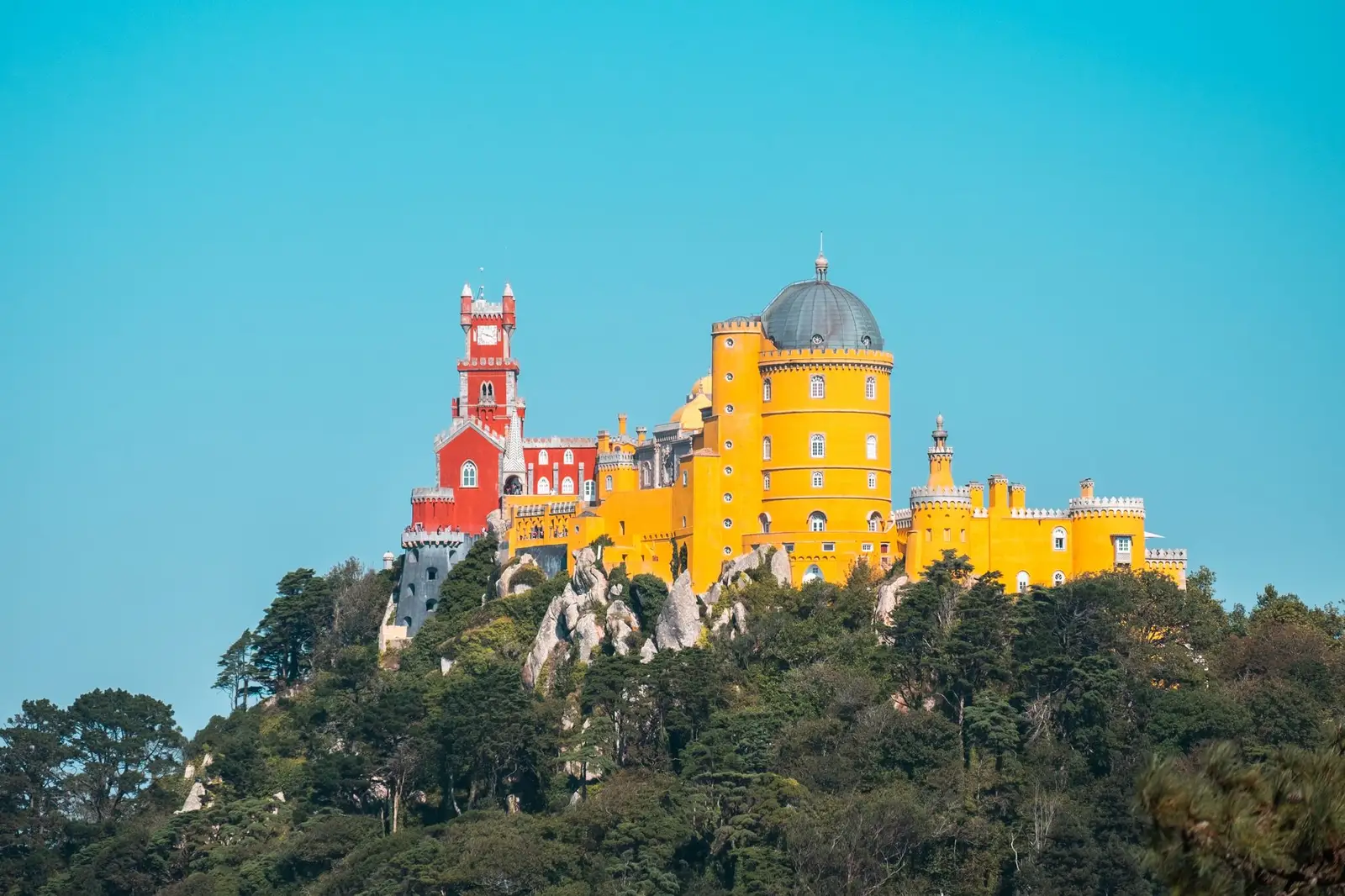 The colorful Pena Palace sits atop a hillside, surrounded by lush greenery under a clear blue sky in Sintra, Portugal.