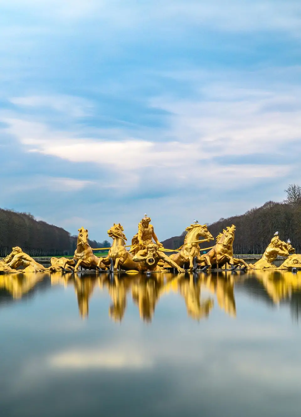 Golden statues in a serene lake at Versailles with trees in the background, France.