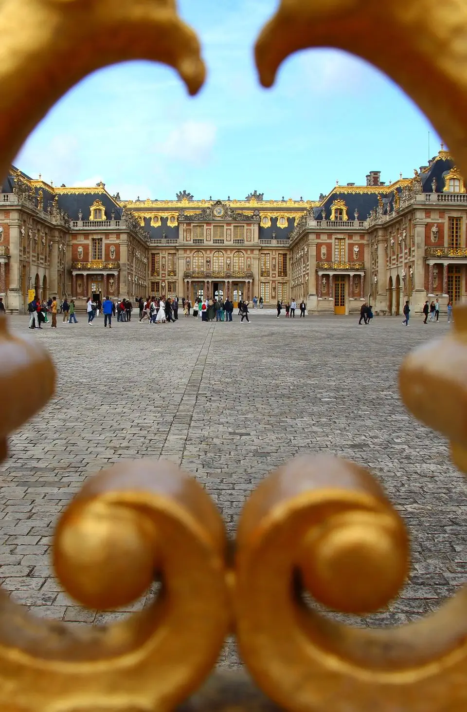 Gilded gate framing the majestic Palace of Versailles courtyard filled with tourists under a bright sky.